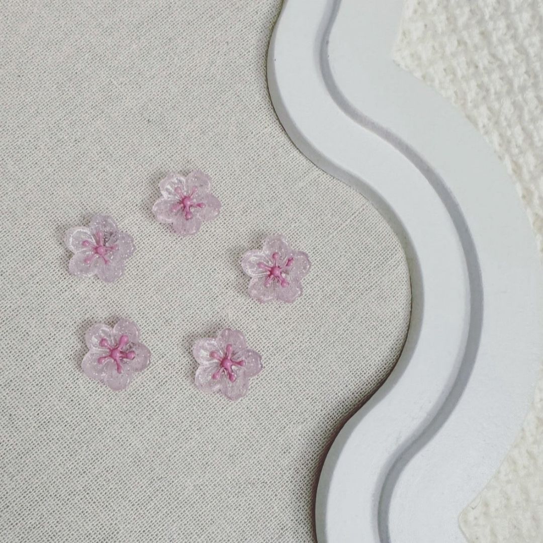 Five translucent pink cherry blossom-shaped magnets with glitter details on a fabric-covered board with a decorative white frame.