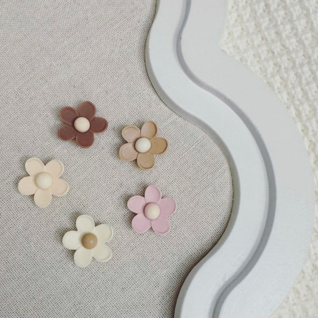 Five pastel and neutral-toned flower-shaped magnets on a fabric-covered board with a decorative white frame.