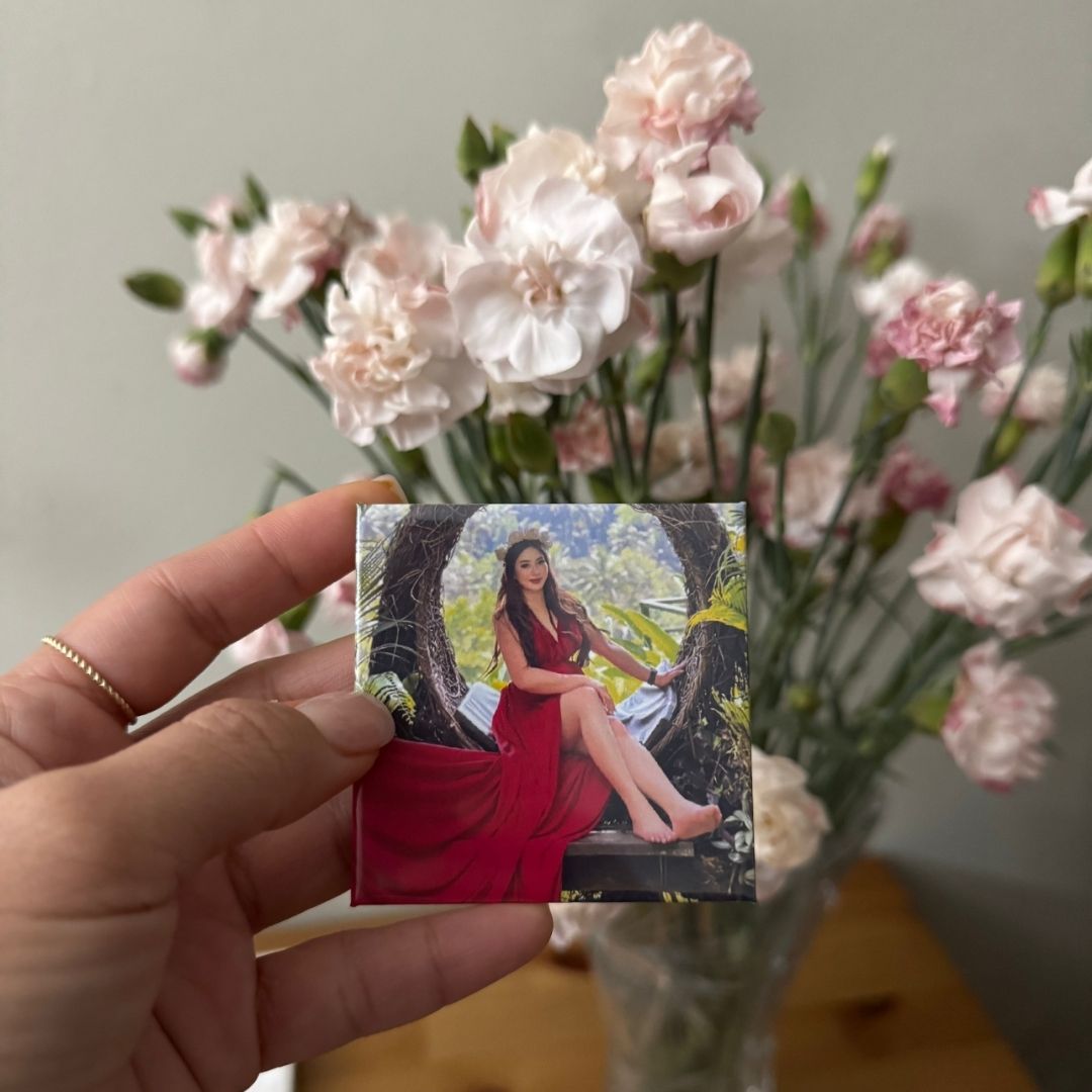 A hand holds a square custom photo magnet print of a woman in a red dress, with pink and white flowers in the background.