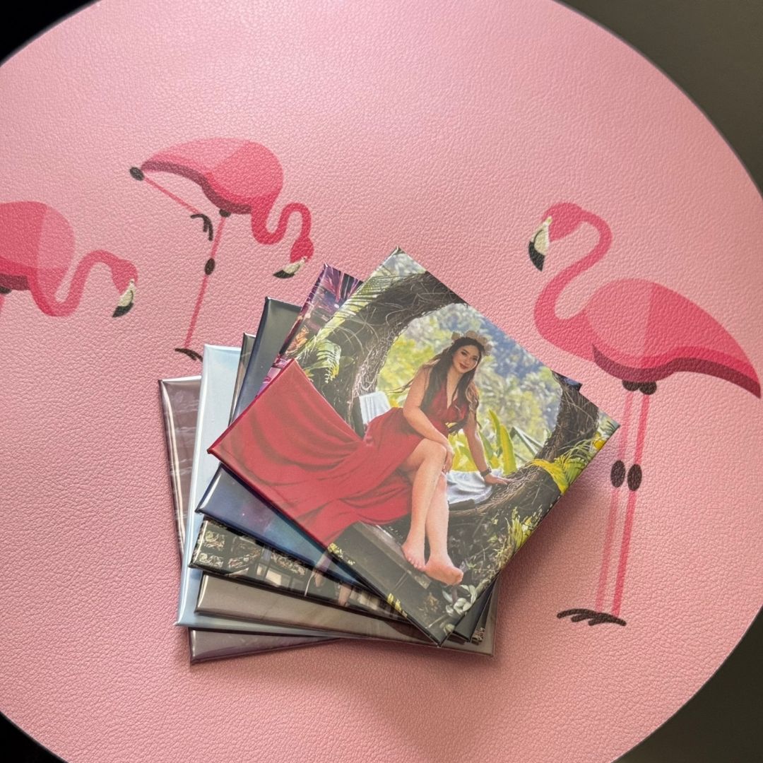 A stack of square custom photo magnet rests on a pink flamingo-patterned surface, with the top print featuring a woman in a red dress.